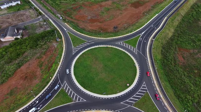 Aerial view shows a circular traffic roundabout. Four roads curve around the green center. Cars move smoothly along the path. Fields surround the roundabout with green and brown patches