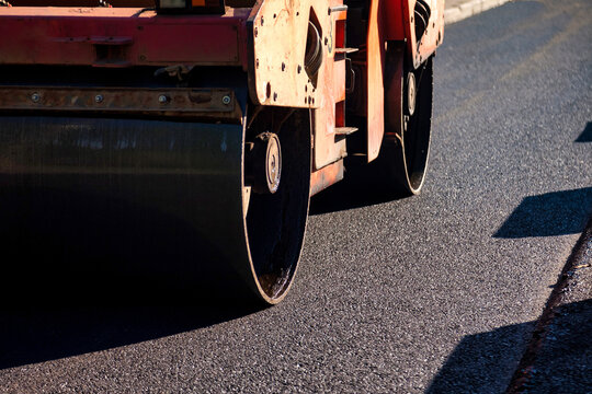 Heavy machinery works on a city street to lay down asphalt concrete. The process involves rolling and smoothing the surface. This work is part of urban development and maintenance.