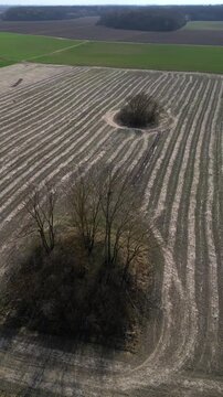 Vertical video. Scenic aerial view flying forward over a rural landscape with interesting patterns in a plowed field where farm machinery has maneuvered around small groves of leafless trees