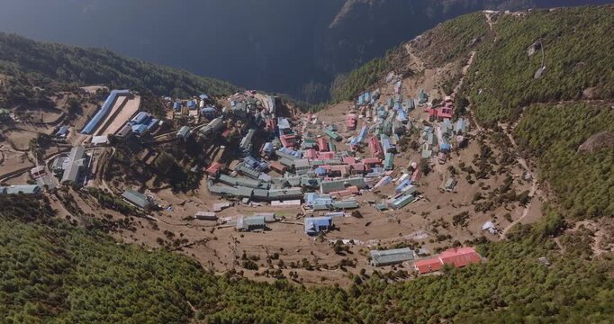 Aerial shot of Namche Bazaar showing its amphitheater-shaped layout with colorful rooftops and surrounded by green forests, rugged hills and snow-capped Himalayan peaks in Nepal.