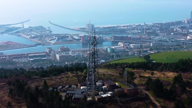 Retreating Aerial View of TV Radio Transmitter Tower High Above Swansea City with Marina in Distance. 5G Infrastructure for Smart Phones and Connectivity.