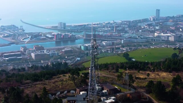 Sideways Aerial View of TV Radio Transmitter Tower High Above Swansea City with Marina in Distance. 5G Infrastructure for Smart Phones and Connectivity.