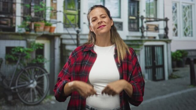 Woman making finger heart gesture with hands, wearing red flannel and white top on a cobblestone street by bicycle and building; playful charm.