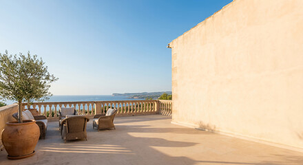 Obraz premium A beautiful terrace with wicker chairs and an olive tree overlooks the sea and coastline under a clear blue sky.