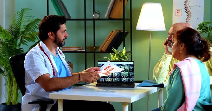 Professional Indian young male doctor explaining organ functions using a graphic figure to Asian senior retired couple patients across a table in a modern clinic; healthcare education concept.