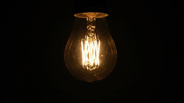 A single incandescent light bulb glows warmly in the darkness, illuminating its intricate filament structure