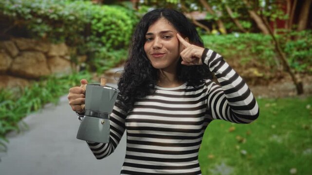 Woman points finger to temple while holding a gray coffeepot on a park pathway by green lawn and stone wall; playful idea.