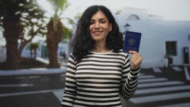 Young brunette woman holds passport up with one hand on a street crosswalk and building behind her; travel confidence.