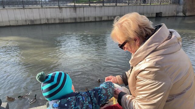 A woman and a young child in winter clothes sit by a city canal. They are throwing bread from a bag to a group of ducks in the water. The scene is bright and sunny.