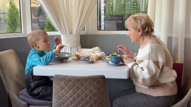 An older woman and a young boy sit at a white cafe table, sharing a meal and warm smiles. They look happy and connected as they eat and talk in the cozy, sunlit room.