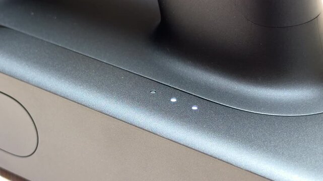 A black electronic device shows a single lit LED light among three small indicator holes. The matte plastic surface reflects light in a clean, modern indoor setting.