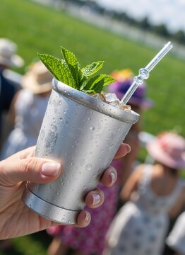 Hand holding frosted silver cup with mint julep cocktail and ice. Cold refreshing drink with condensation drops. Blurred background of people at outdoor derby event