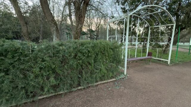 A peaceful wooden bench sits tucked inside a white metal frame in Vorontsov Park. Dense green hedges and tall trees surround the area, creating a quiet and cozy spot for morning reflection.