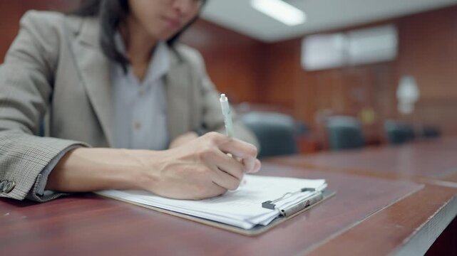 Professional woman writes notes at wooden desk. Focused expression shows concentration during work. Handheld pen and clipboard capture moment of preparation. Interior lighting highlights desk surface