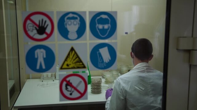 Laboratory safety signs on glass door with researcher working inside, illustrating protective equipment requirements and hazard warnings in controlled environment