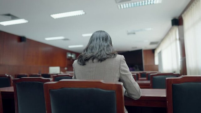 Woman sits in courtroom, facing judge's bench. Grey hair flows softly, draped over shoulders. Wooden chairs, tables, and polished floors. Rows of empty seats behind her, quiet and still
