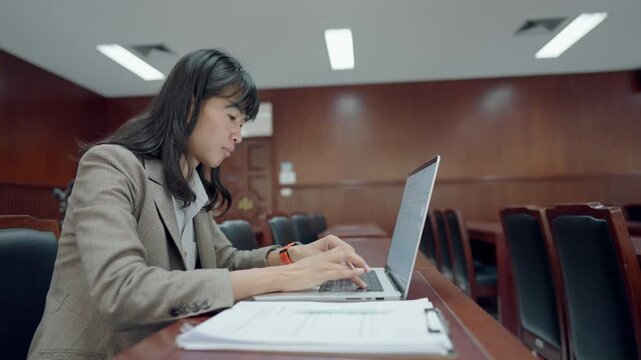 Professional woman uses laptop in courtroom setting. She types actively, focused on task. Wooden benches surround her, formal environment. Her attire suggests legal or administrative role