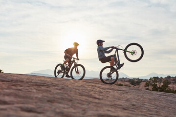 Fototapeta premium Nature, men and extreme sport with balance on mountain bike for action stunt, adventure and space. Sunlight, bicycle and friends with wheelie on rocky terrain for adrenaline, freestyle trick or hobby