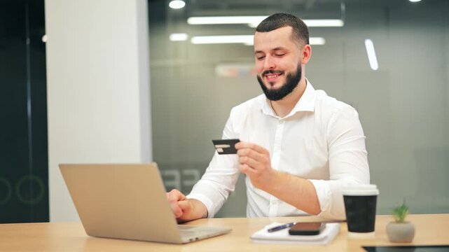 Joyful bearded man in white shirt making secure digital payment with credit card and laptop. Successful male executive enjoying online shopping or banking at office desk.