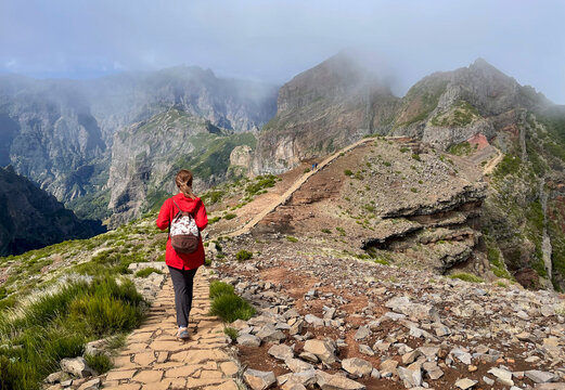 A hiker in a red jacket follows a stone-paved trail along the high volcanic ridges of Pico do Arieiro, surrounded by jagged peaks and drifting mountain mist.