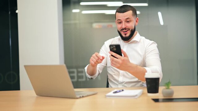 Man receives positive news on mobile device, demonstrating initial surprise followed by pure happiness and excitement. Celebrating personal achievement at modern office desk, satisfied with results.