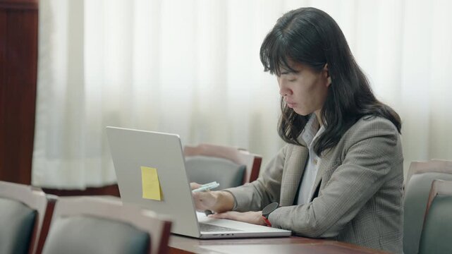 Professional woman types on laptop in meeting room. She wears business jacket and focuses on screen. Yellow sticky note attached to her laptop. Soft light filters through window blinds