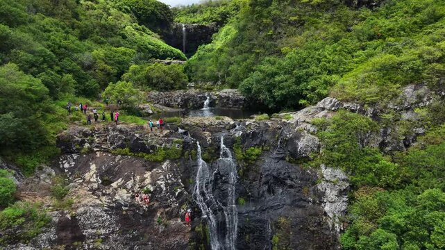Keyframe 1: Green mountains frame a small waterfall. Hikers walk on rocky terrain. They smile, enjoying nature's view. Sunlight glints on flowing water. Lush trees surround the area