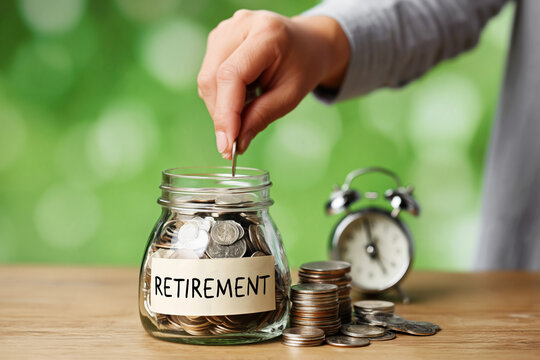 A persons hand carefully places a shining coin into a glass jar labeled Retirement The jar is full of money sitting on a wooden table with stacked coi