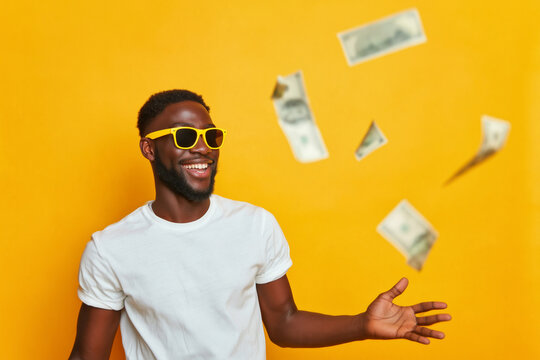 Joyful young Black man in bright yellow sunglasses broadly smiling as money bills rain down around him against a vivid yellow backdrop conveying happi