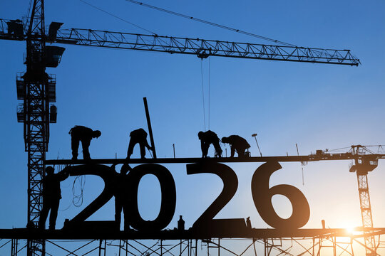 Silhouetted construction workers are diligently assembling the huge metallic numbers 2026 on a high scaffolding platform with towering industrial cran