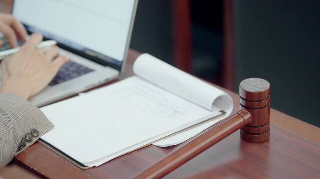 Courtroom setting with wooden gavel and open notebook. Hand writes notes while using laptop. Professional attire suggests legal or judicial context. Scene focuses on preparation or review of documents