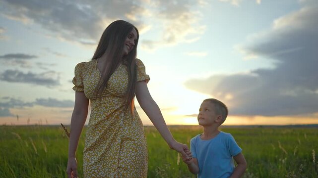 Mother and son walk in green field at sunset holding hands. Happy family moves through grass during summer evening. Mother and son enjoy nature outdoors. Family goes forward in sunset field.