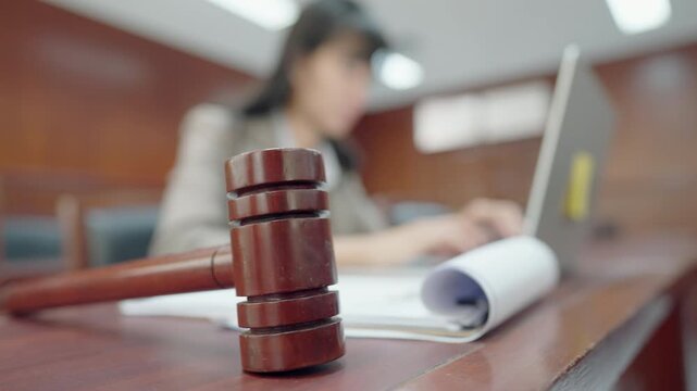 Wooden gavel rests on polished desk. Blurry figure works on laptop in background. Courtroom setting with official tones. Focus remains on gavel and documents. Professional atmosphere captured sharply