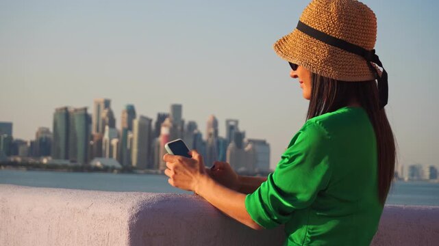 Woman tourist wear green dress and straw hat checks her using smartphone navigate at the Doha Corniche waterfront . The West Bay skyline and Persian Gulf create a modern backdrop