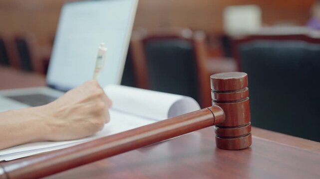 Wooden gavel rests on desk beside laptop. Hand writes notes beside open notebook. Courtroom setting with blurred background chairs. Lawyer works with digital device and paper records