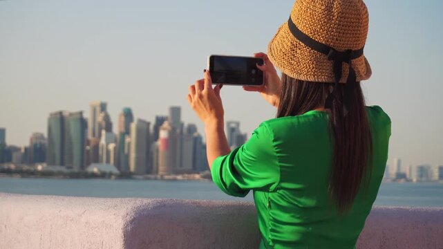 Tourist in green dress taking a smartphone photo of the West Bay skyline from the Doha Corniche. Bright sunny day with the Persian Gulf in the background