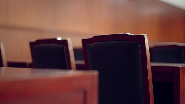 Rows of dark chairs line a courtroom's wooden floor. Wooden desks and benches stand empty in the room. The space appears quiet and still