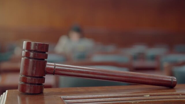 Wooden gavel rests on courtroom table. Blurred judge sits behind gavel. Rows of empty seats stretch into background