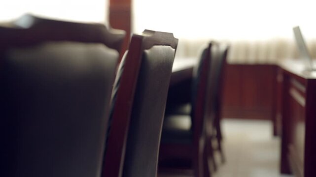 Rows of dark chairs line a long wooden table. Blurred background hints at a conference or courtroom. Soft focus creates an air of quiet anticipation. Dark wood surfaces gleam under subdued lighting