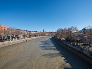 Wide angle view of the Mtkvari River flowing through Tbilisi, Georgia, with the Holy Trinity Cathedral (Sameba) visible on the horizon under a clear blue sky in winter. © Microscope