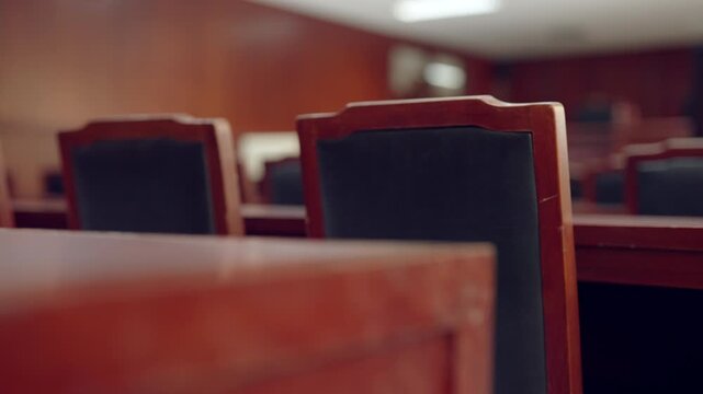 Empty courtroom chairs in polished wood. Rows of dark-backed seats face a distant bench. Soft focus emphasizes the quiet, solemn atmosphere. Light reflects off polished surfaces with warm tones