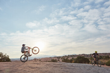 Fototapeta premium Man, mountain bike and wheelie in summer with friends, sky and extreme sports for trick with balance. People, bicycle and sunshine with ride, exercise and outdoor on hills with space in countryside
