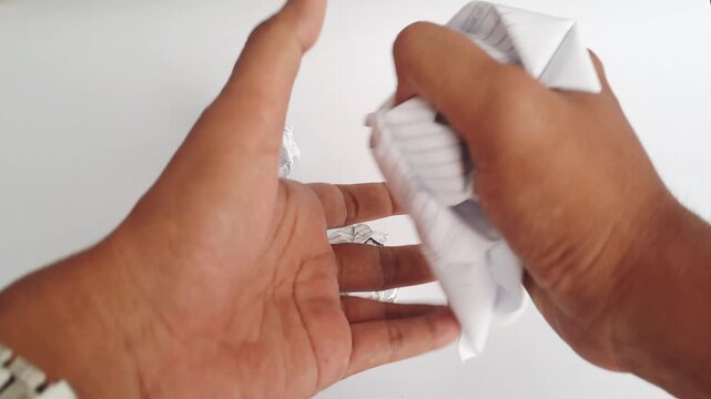 Close-up of an Asian man's hands crumpling a piece of white paper and throwing it away in anger. A minimalist horizontal shot depicting frustration, failure, creative block, and stress at work.