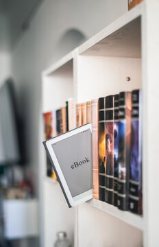 Ebook reader placed between paper books on shelf. Digital reading meets traditional library, modern technology and education concept in cozy home interior. Vertical photo