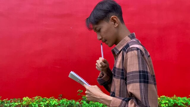 Young man in shirt ponders ideas, jotting notes in notebook against vivid red backdrop