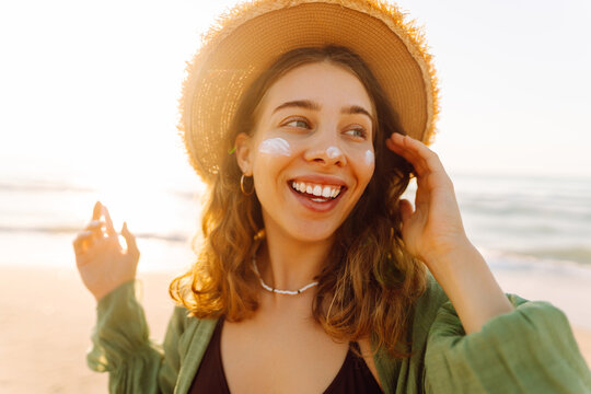 Close-up of a young woman applying sunscreen to her face on a sunny beach. A happy woman enjoys the warm sun and relaxes by the sea. Concepts: relaxation, protection, care.