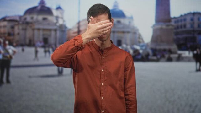 Man young covering face with right hand in a busy street plaza, wearing orange shirt and glasses while standing before a domed church and obelisk; shame solitude.