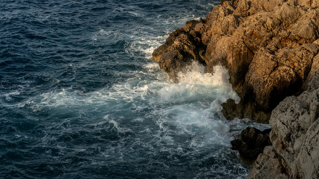 Waves Crashing Against Sunlit Rocky Coastline. Powerful waves crash against rugged coastal rocks under warm natural light. 