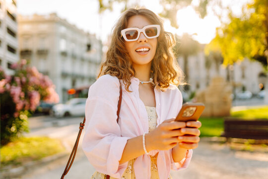 A tourist in a dress, using her phone, enjoys a stroll in a sunny park. Beautiful woman strolls outdoors with phone at sunset. Concept of tourism, walking, technology.