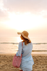 A sweet woman in a hat and carrying a red bag strolls along the beach at sunset. Young woman enjoys...